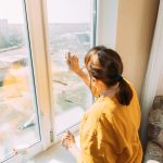 Woman Of Fifty In Yellow Sweater And Jeans Washes Dusty Window In Apartment. 50 Year Old Woman